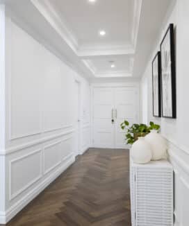 Elegant hallway featuring Intrim SK60 skirting, complemented by white walls and herringbone timber flooring. A console table with sculptural vases and greenery sits beneath framed monochrome artwork, leading to double doors that anchor the classic French Provincial interior design.