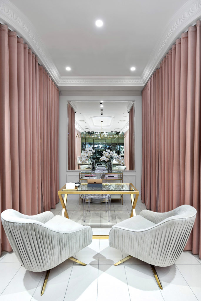 A plush space in a retail shop for customers to sit. A white tile floor is surrounded on both sides with pale pink curtains and the back wall features floor to ceiling wainscoting with a large decorative mirror reflecting back into the rest of the shop.