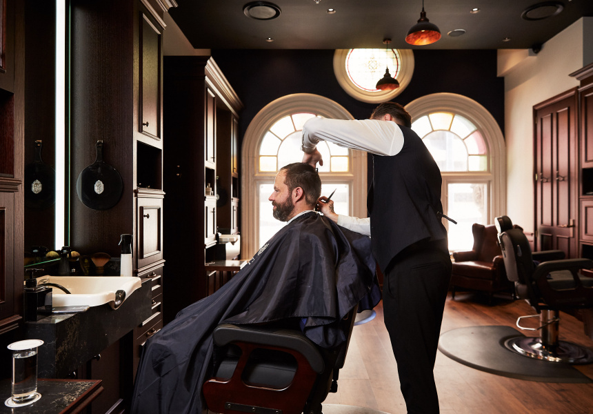 An upscale barber's featuring dark walling framed with curved windows and custom curved architraves, dark wooden flooring, and stylish seating. A barber is trimming the hair of a male customer.