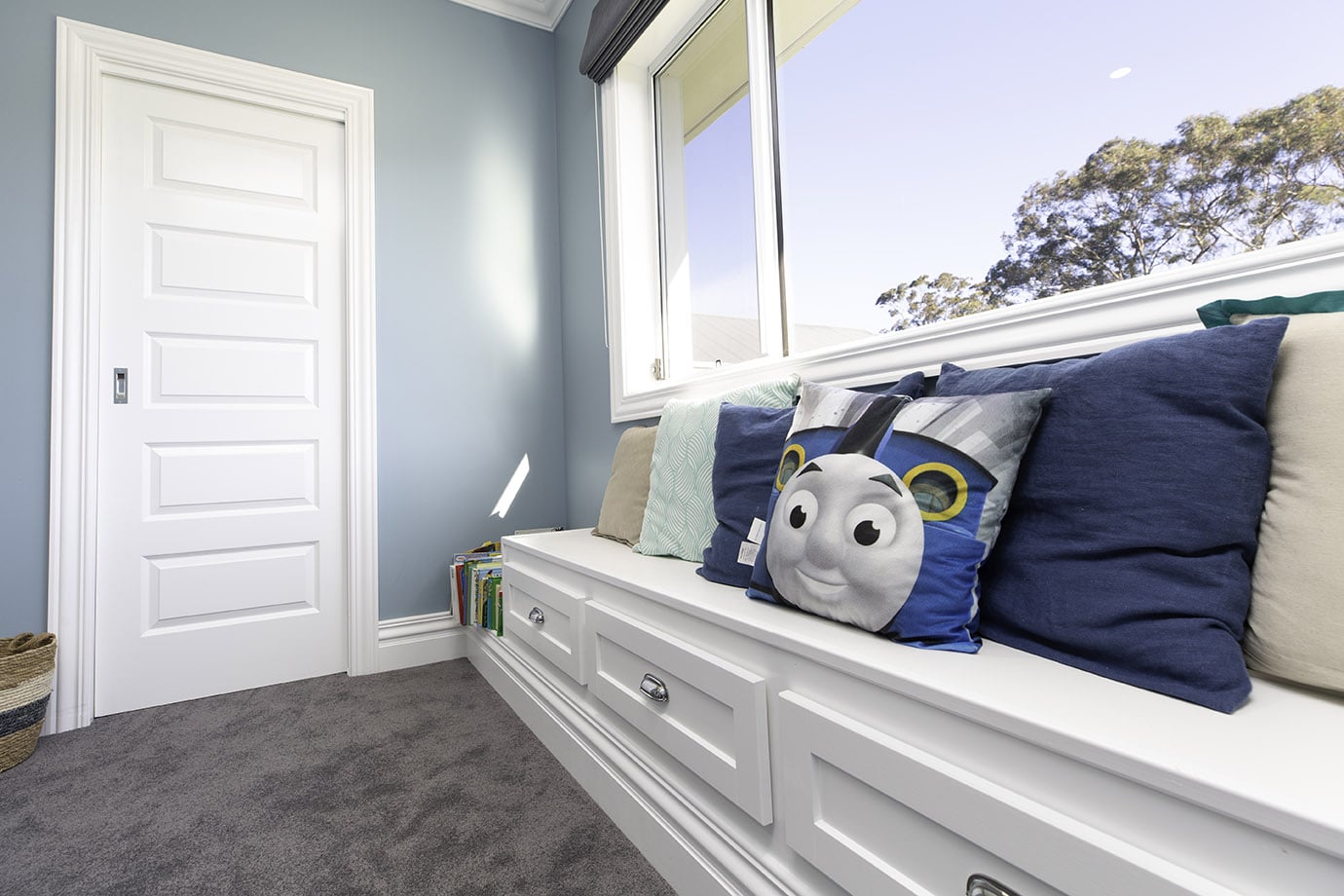 A boy's bedroom with bench seat underneath a window, painted white, and Intrim SK460 skirting and architrave framing a white door. Grey carpet lines the floors and a Thomas the Tank Engine pillow sits on the bench seat.