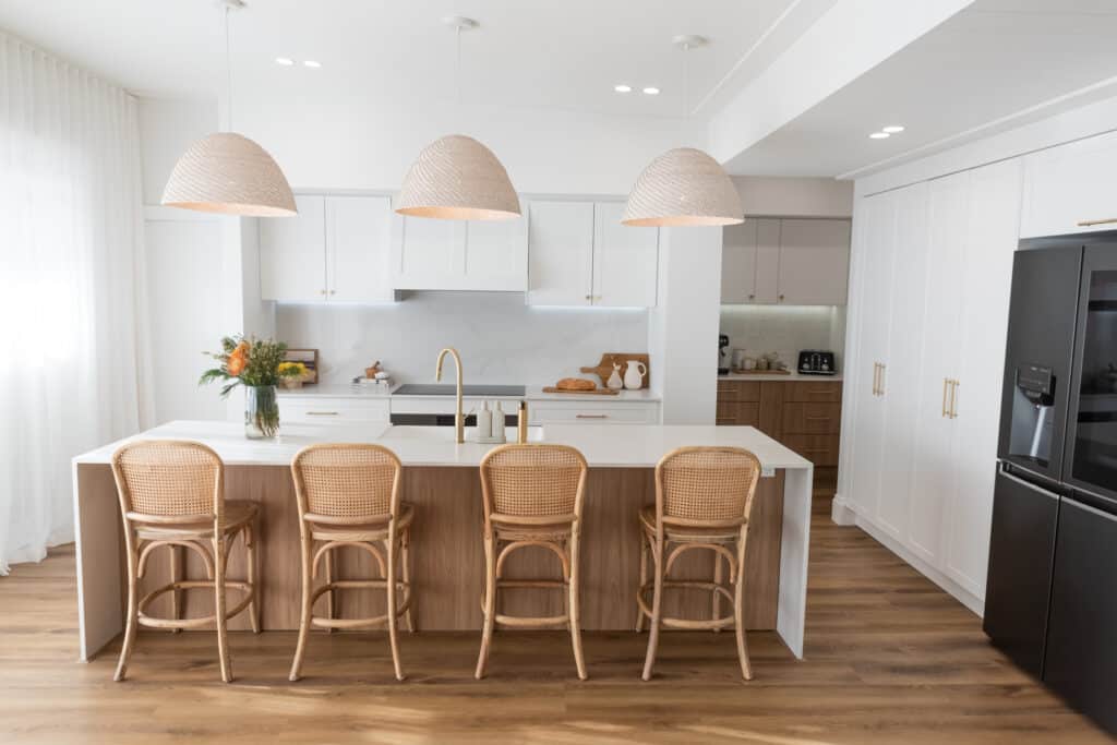 Beautiful kitchen in Scandinavian Coastal style featuring a large white kitchen island, rattan-backed chairs, and three white, domed pendant lights