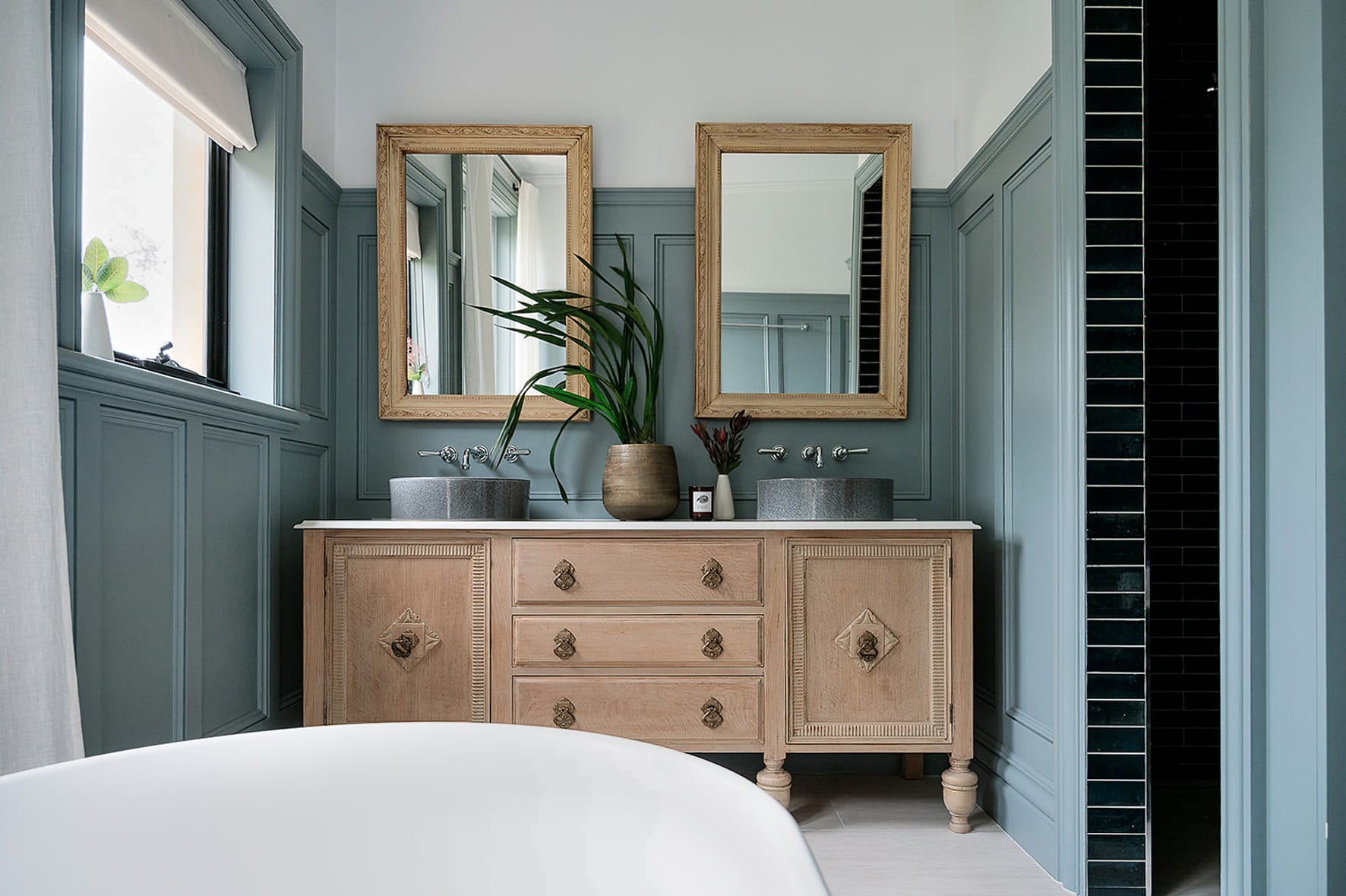 Traditional bathroom featuring half-height wall panelling painted in muted blue-green, paired with classic mouldings, twin framed mirrors and a timber vanity for a timeless look