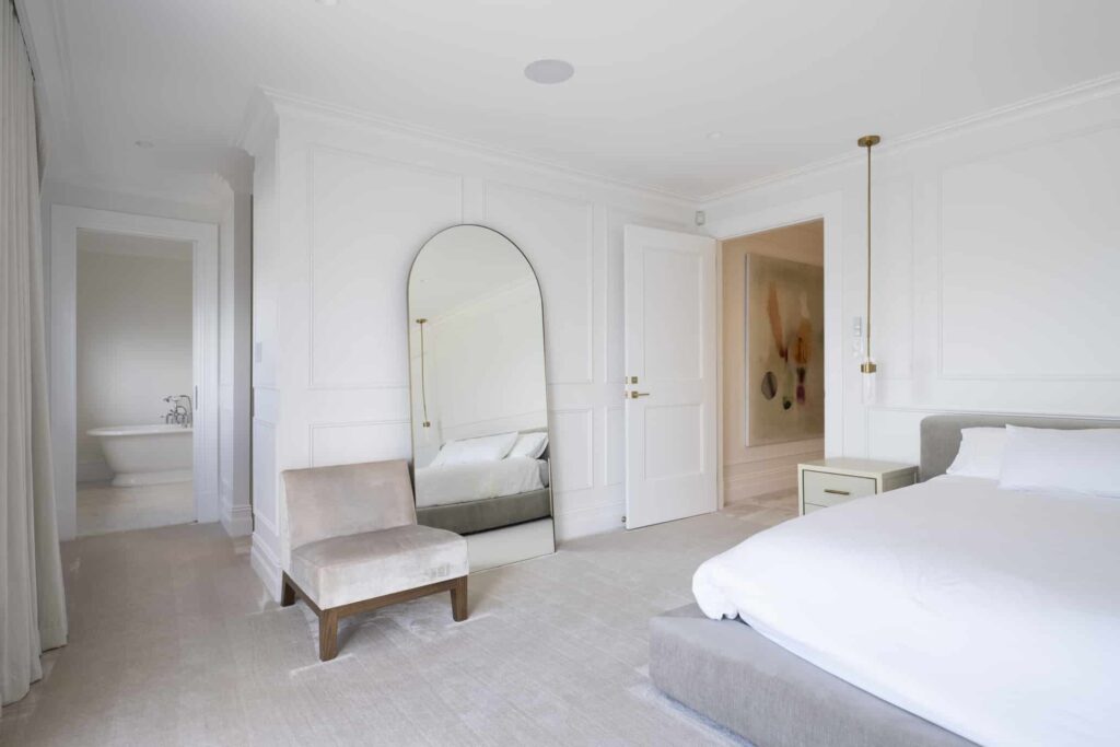 Modern Hamptons-style bedroom showcasing white wall panelling and light wooden flooring with a large white bed, feature mirror, and accent chair