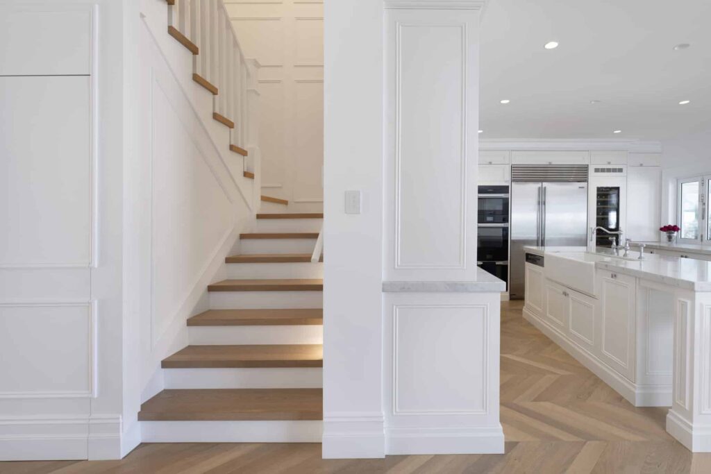 Modern Hamptons-style kitchen with contemporary white cabinetry, marble countertop, herringbone flooring, and a wooden staircase
