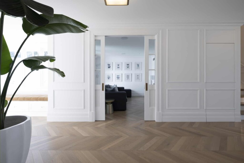 Airy Modern Hamptons-style living space showcasing white panelled wall and wooden herringbone flooring with a house plant in the foreground
