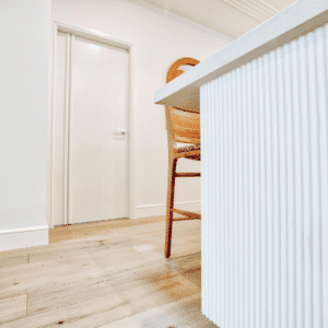 Close up of a Scandinavian Coastal-style kitchen island featuring vertical white timber panelling against a light wooden floor with a wooden dining chair