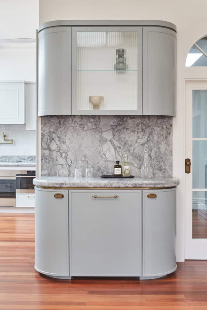 Curved kitchen bar cabinetry in muted grey tones with dramatic marble backsplash—mouldings enhance the bespoke craftsmanship and visual flow.