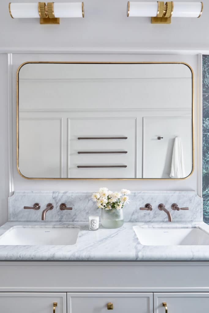 Dual-sink vanity with marble countertops, brass fixtures, and a gold-framed mirror—mouldings elevate the bathroom’s classic luxury aesthetic.