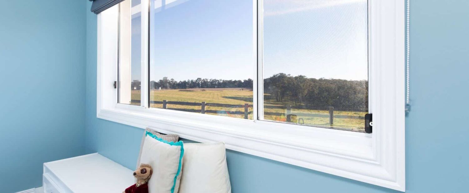 Angled close-up of crisp white architrave moulding around a window through which a green field and clear sky can be seen. Blue walls and cream cushions feature in the foreground.