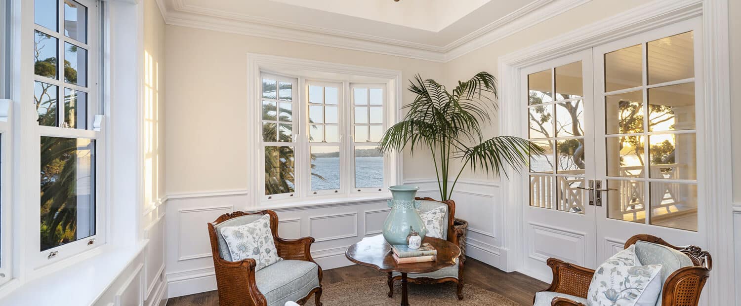 Coastal-inspired sunroom with ocean views, wicker armchairs with light blue cushions, round wooden table styled with books and a green vase, potted palm, an elegant chandelier, and Intrim's premium decorative timber mouldings.