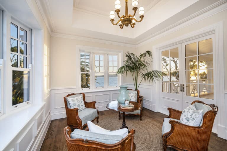 Coastal-inspired sunroom with ocean views, wicker armchairs with light blue cushions, round wooden table styled with books and a green vase, potted palm, an elegant chandelier, and Intrim's premium decorative timber mouldings.