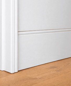 A close up of a soft, curved flush finish skirting board and coordinated curved door jamb, painted white against timber flooring.
