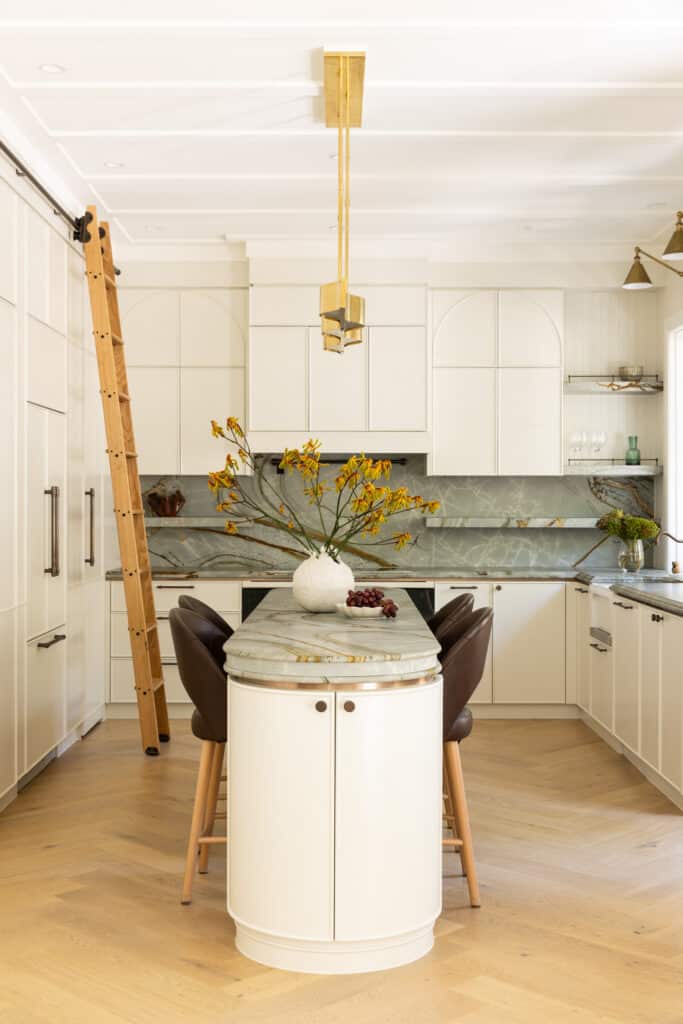Cream shaker kitchen with a curved stone-topped island, brown leather counter stools, green-veined splashback and a sculptural brass pendant.