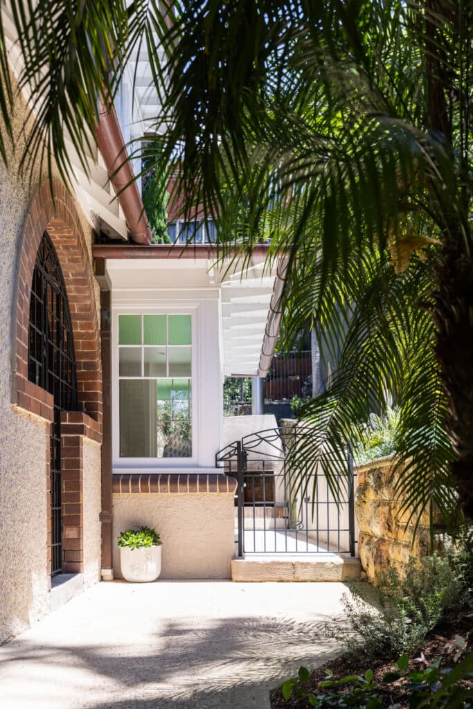 Exterior of a heritage Neutral Bay home with a rendered brick archway, sandstone retaining wall and established palms beside a white-framed timber window.