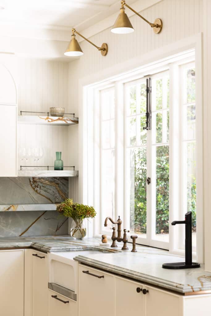 Kitchen corner with twin brass wall lights, open shelving, a veined stone splashback and aged-brass tapware beside full-height French windows finished with Intrim SK498 architrave.