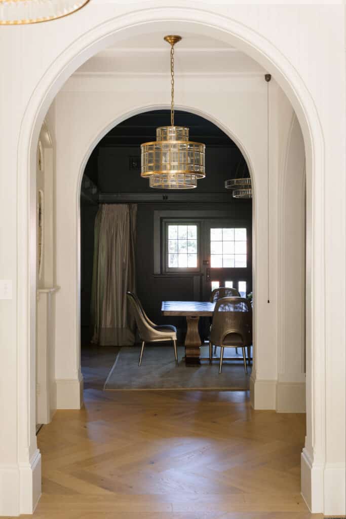 View through a white archway into a moody green formal dining room with a timber trestle table, woven chairs and a brass drum pendant, framed by Intrim SK498 custom curved architraves and skirting.