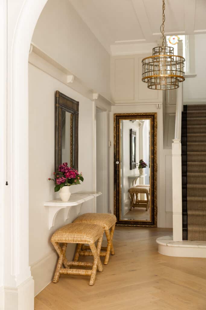 Light-filled entry hall with a marble bracket console, woven X-base stools, gilt floor mirror and brass lantern beside a carpeted staircase, finished with Intrim SK498 skirting