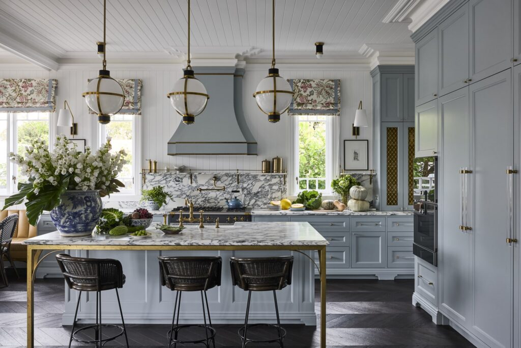 Hamptons-style kitchen with decorative wall panelling, VJ ceiling lining, and elegant cornice detailing in a soft blue palette, elevated by marble and brass accents.
