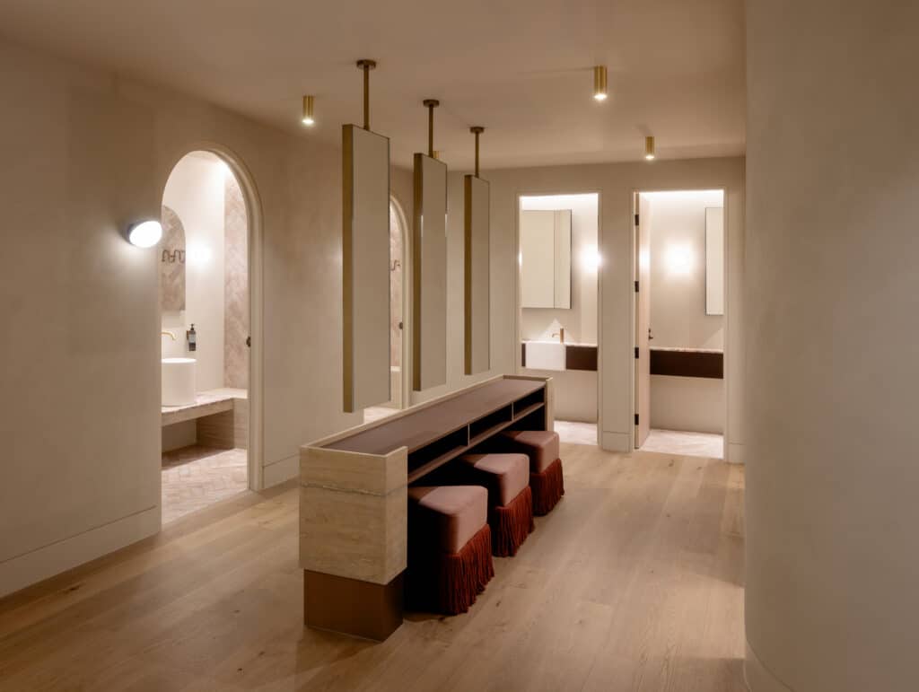End-of-trip amenities room at Zagame's Place Hawthorn by Studio Arcka, with a travertine stone bench, burgundy velvet stools, tall hanging mirrors on brass rods, and arched doorways leading to bathroom vanity areas with Intrim DJ800 integrated door jambs