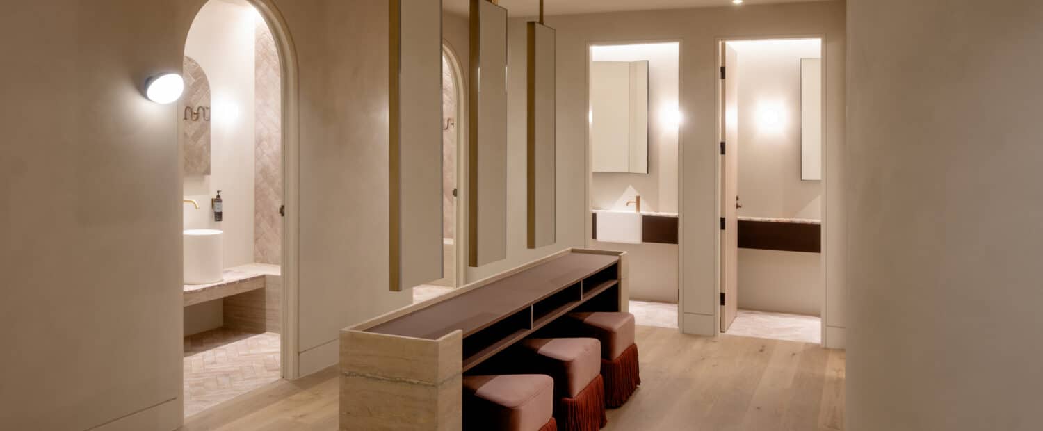End-of-trip amenities room at Zagame's Place Hawthorn by Studio Arcka, with a travertine stone bench, burgundy velvet stools, tall hanging mirrors on brass rods, and arched doorways leading to bathroom vanity areas with Intrim DJ800 integrated door jambs