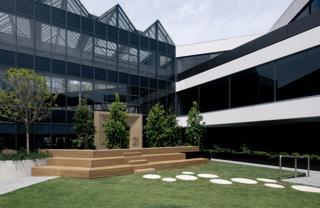 Exterior courtyard of Zagame's Place at 2 Luton Lane Hawthorn, showing the building's distinctive sawtooth glass roofline, a central timber deck platform, manicured lawns with circular stepping stones, and landscaping by Jack Merlo
