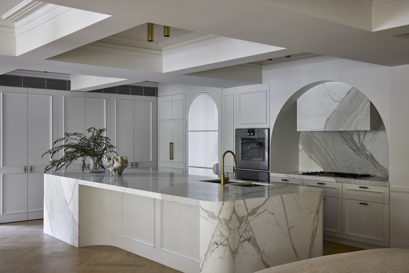 Luxury kitchen with a book-matched marble island, arched marble splashback and range hood, white shaker joinery and brass tapware on herringbone floors