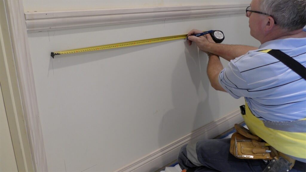 Installer measuring between a dado rail and skirting board with a tape measure to mark out wall panel mouldings.