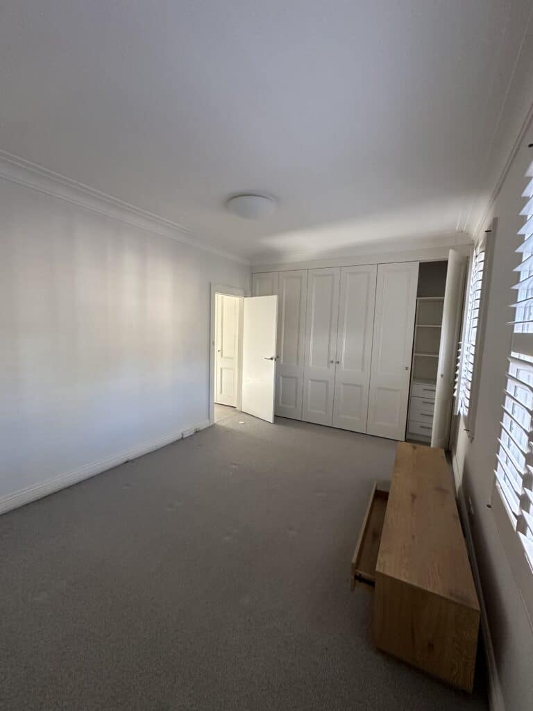 A white bedroom with white walls, grey carpet, built-in panelled wardrobes and plantation shutters on the windows.