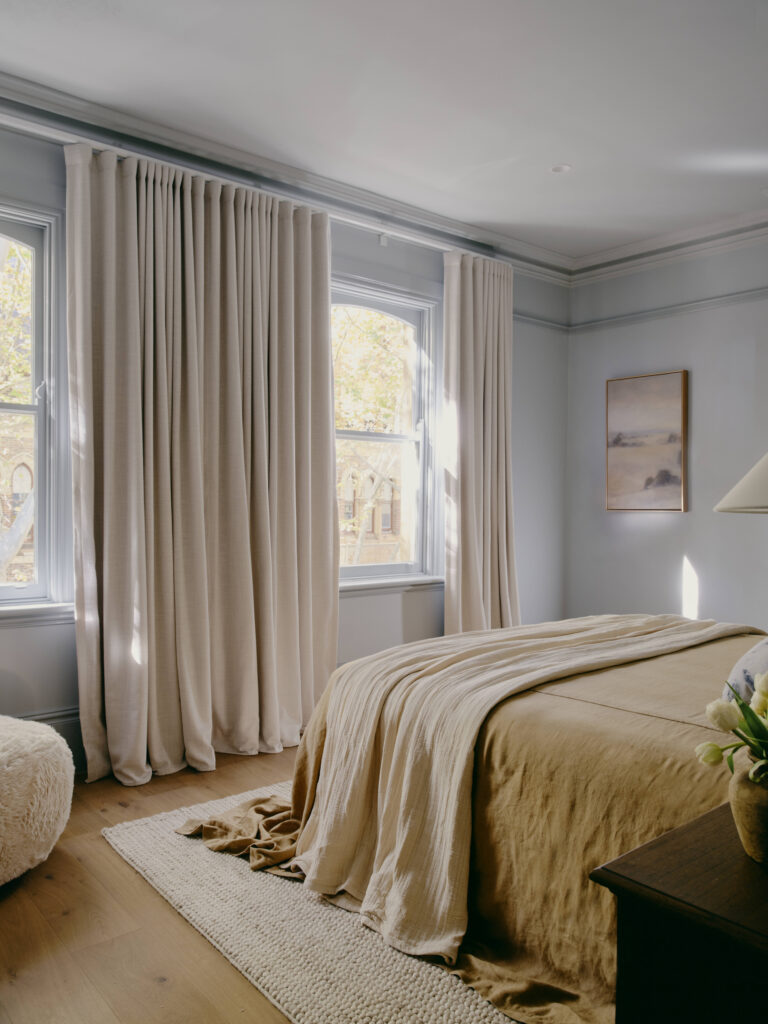 Primary bedroom with pale blue walls, a picture rail and cornice, floor-length linen curtains over two sash windows and a bed in ochre and cream linen.