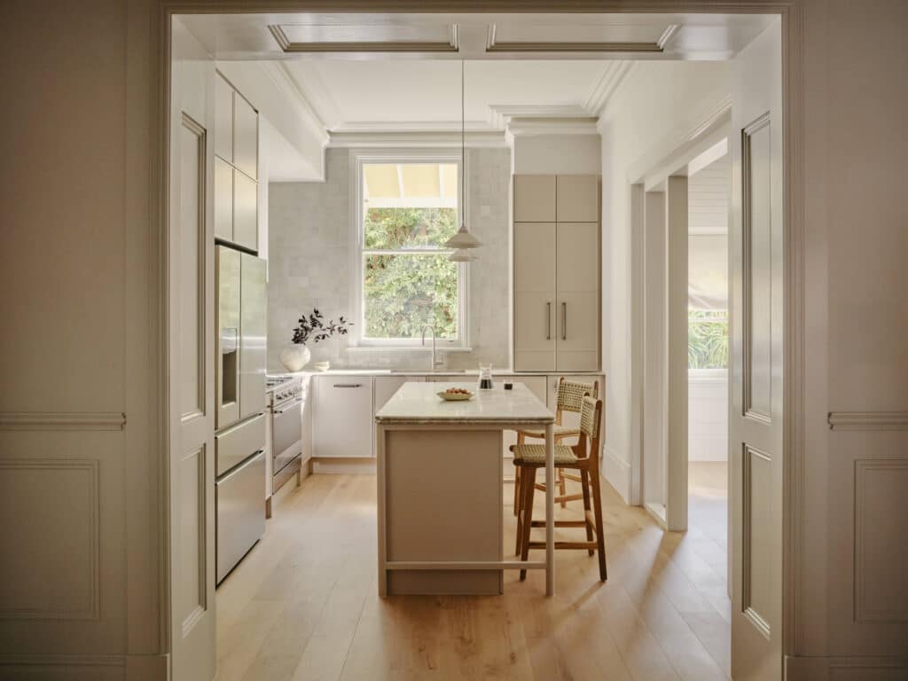 Terrace kitchen seen through a wide panelled opening, with a stone-topped island, woven timber stools, full-height cabinetry and detailed ceiling cornices.