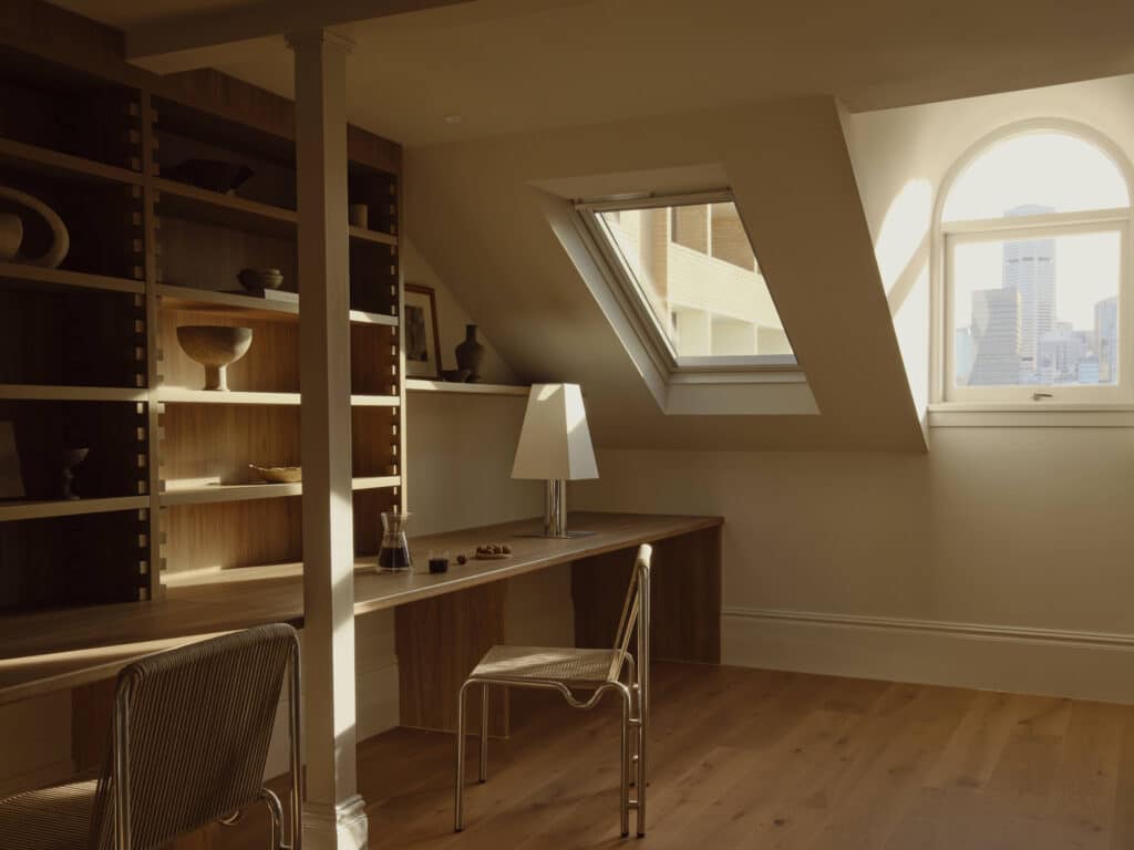 Attic library and study with timber shelving, a long desk for two, a skylight in the sloped ceiling and an arched window looking out to the city skyline.